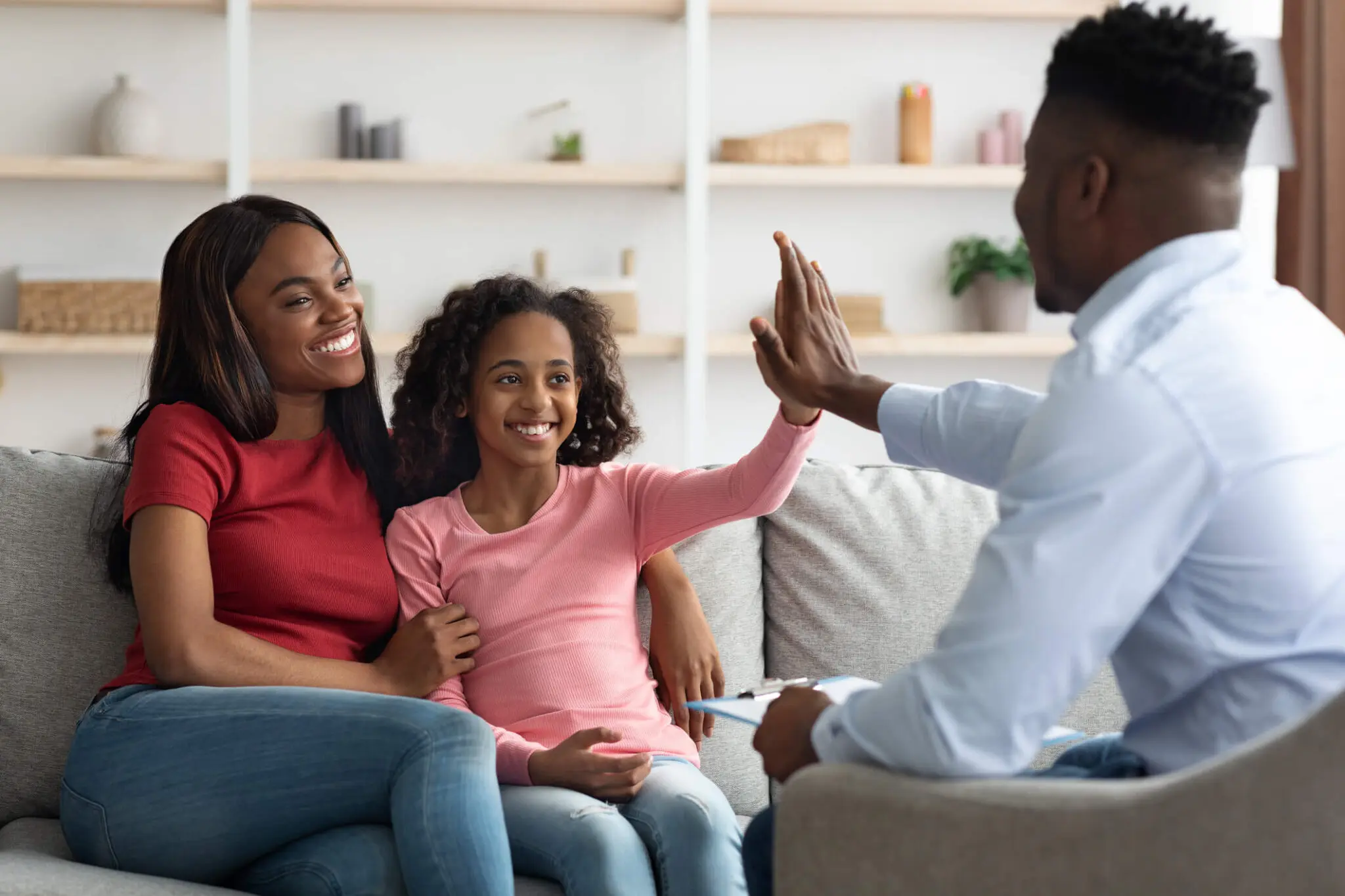A man high-fives a smiling girl sitting on a couch with a woman. Shelves with books and decorations are in the background.