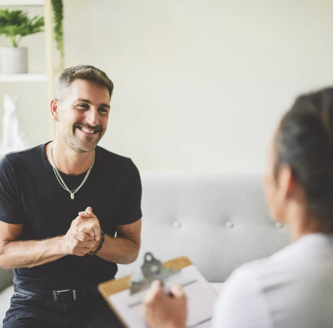 Person wearing a black shirt smiles while engaging in conversation with another person taking notes on a clipboard.