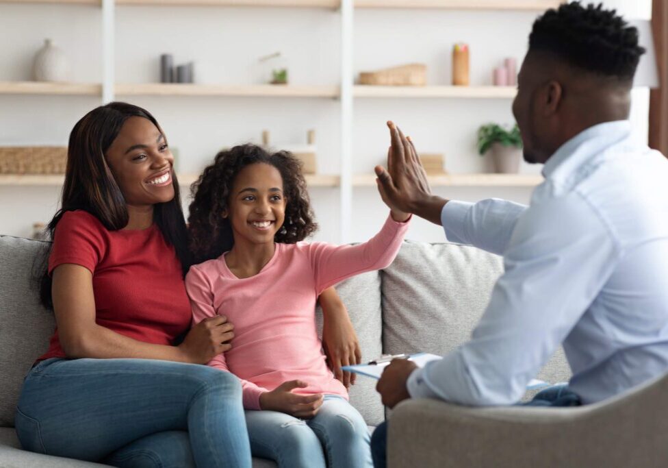 A man high-fives a smiling girl sitting on a couch with a woman. Shelves with books and decorations are in the background.